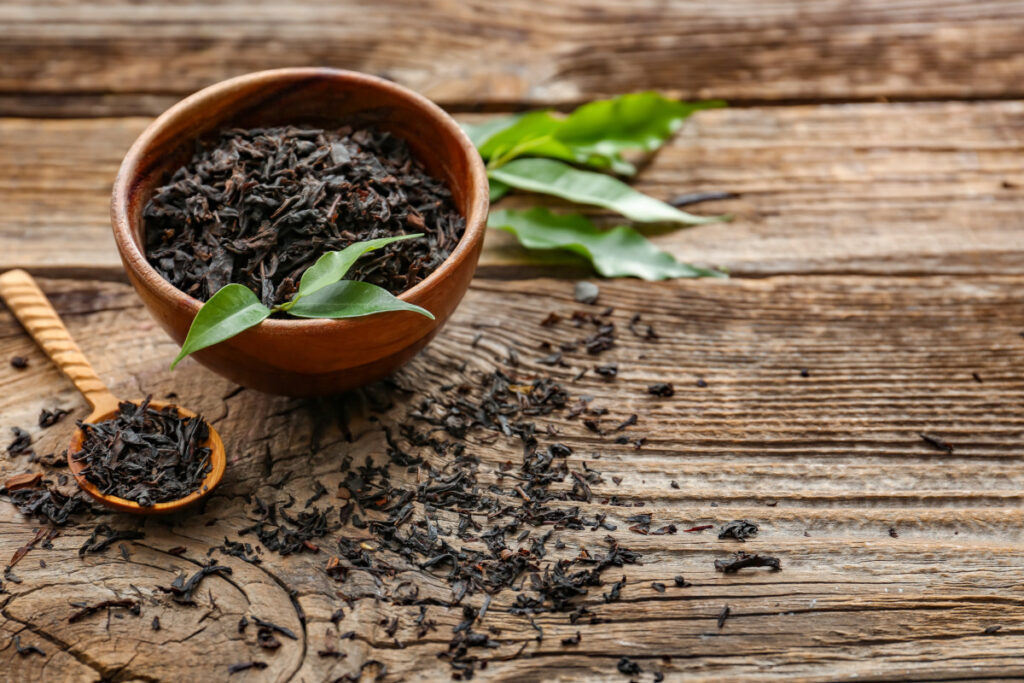 dry black tea leaves in bowl and spoon on wooden background